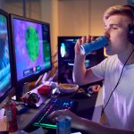 Teenage Boy Drinking Caffeine Energy Drink Gaming At Home Using Dual Computer Screens At Night