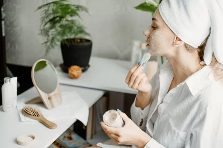 Young woman in white towel chilling in bedroom and making clay facial mask near mirror. Girl doing