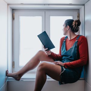 Young woman sitting next to a window reading a book during a bright day, reflexion and self care