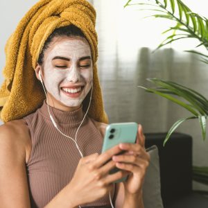 Young woman using mobile smartphone while having skin care spa day at home